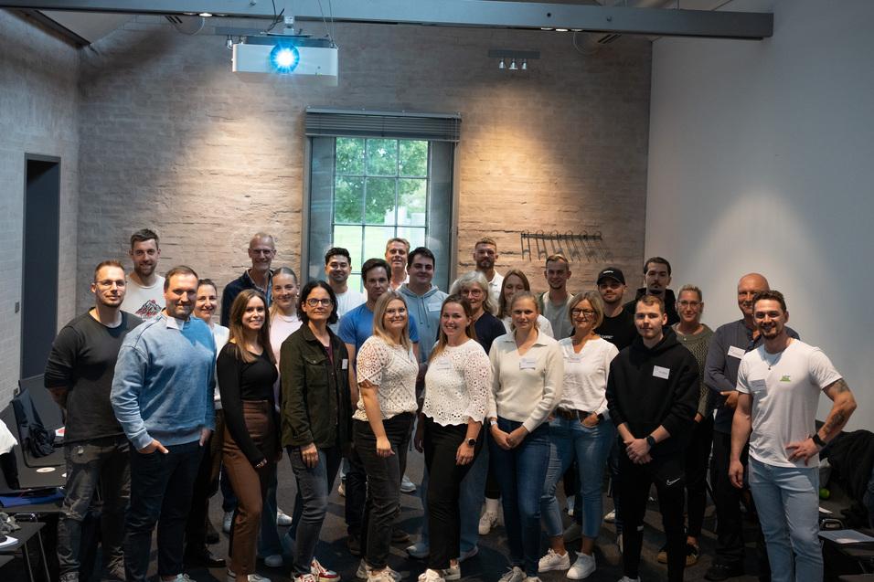 Gruppenshot von 24 Personen in einem modernen Raum mit Fenster und Projektor, alle in l&auml;ssiger Kleidung.