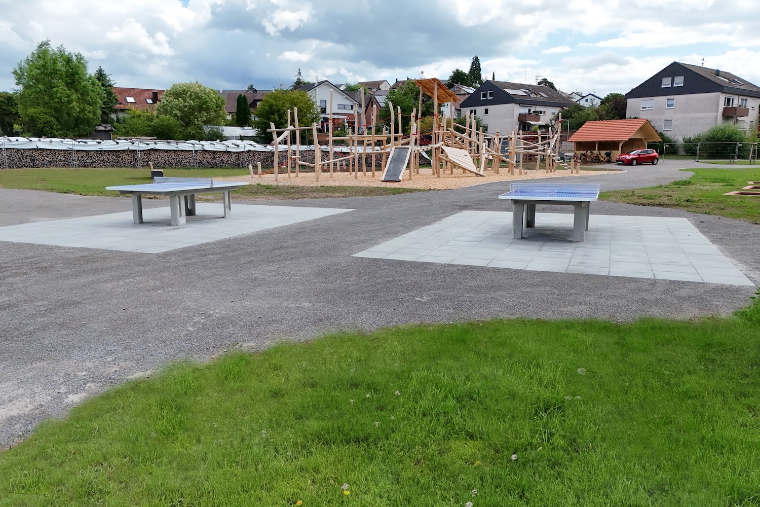 Zwei Tischtennisplatten auf einem Platz, im Hintergrund ein Abenteuerspielplatz unter bew&ouml;lkten Himmel.