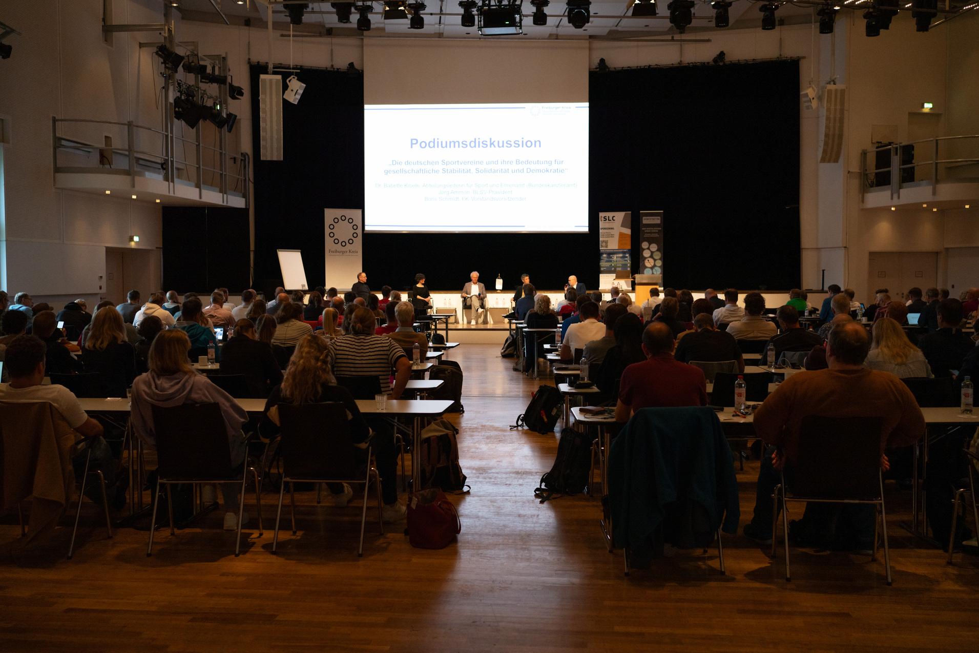 Podiumsdiskussion mit mehreren Rednern auf der B&uuml;hne vor Publikum in einem lichtdurchfluteten Veranstaltungsraum.