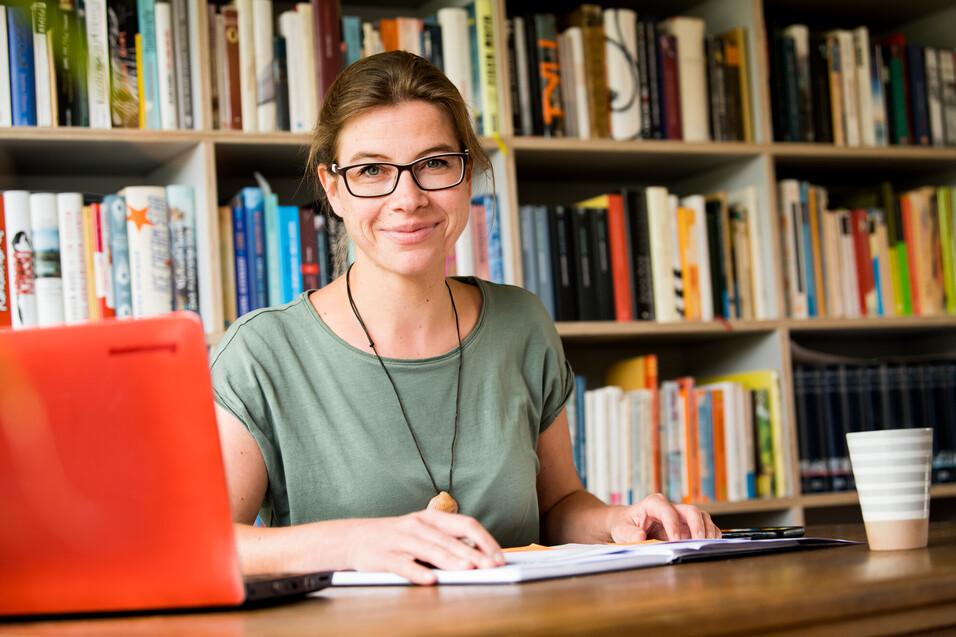 Frau mit Brille sitzt an einem Tisch, umgeben von B&uuml;chern, mit Laptop und Notizen vor sich.