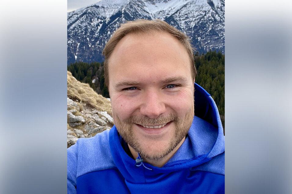 Mann mit blauer Jacke posiert lächelnd vor schneebedeckten Bergen und bewaldeter Landschaft. Wolkiger Himmel im Hintergrund.