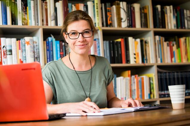 Frau mit Brille sitzt an einem Tisch, umgeben von B&uuml;chern, mit Laptop und Notizen vor sich.