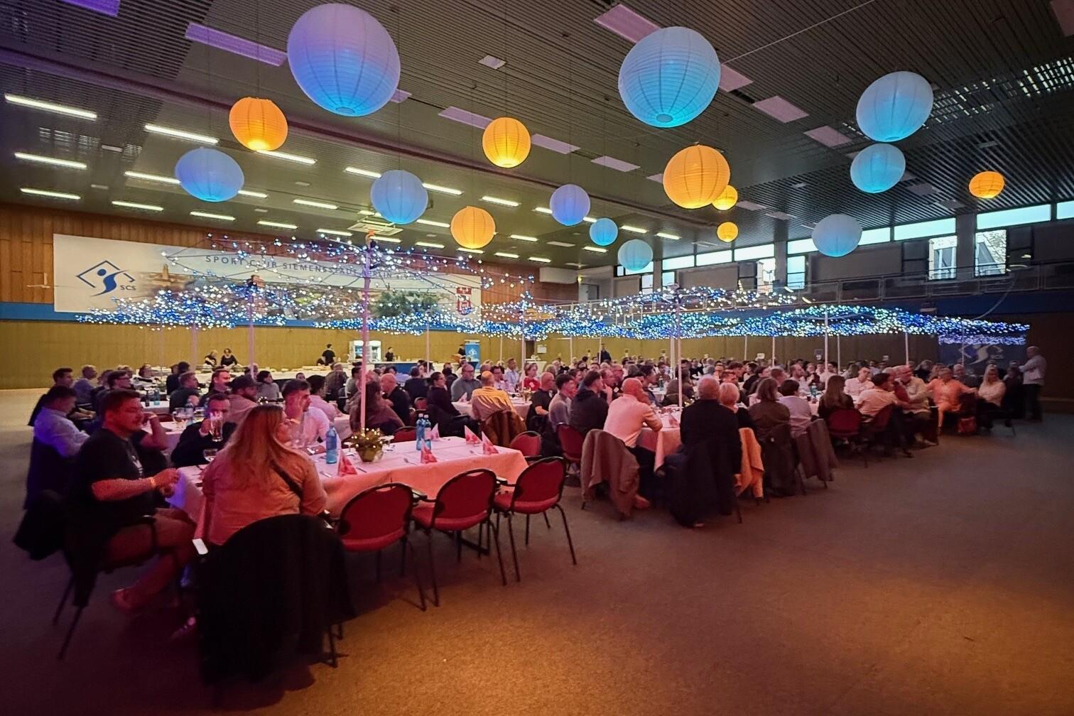 Festliche Veranstaltung mit bunt dekorierten Laternen und Tischgedecken in einer Sporthalle, viele G&auml;ste sind zu sehen.