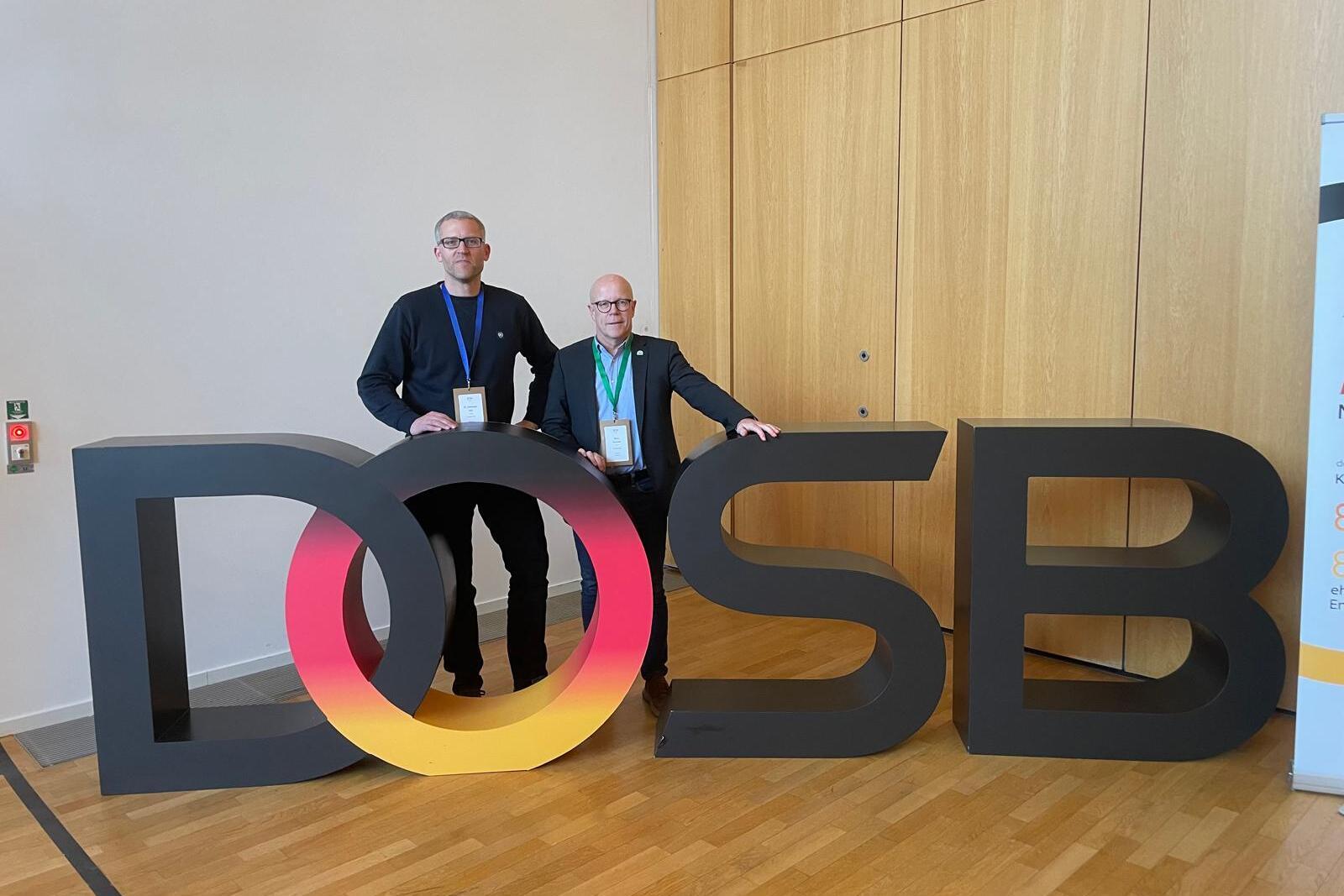 Zwei Personen posieren neben gro&szlig;en, bunten Buchstaben "DOSB" in einem Konferenzraum.