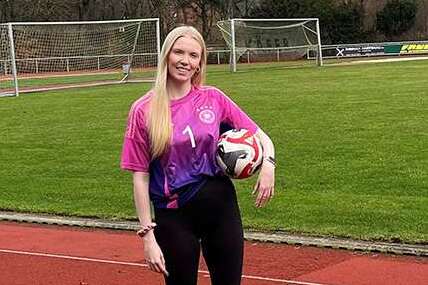 Junge Frau in pink-blauem Fu&szlig;balltrikot mit Ball steht auf einer Laufbahn vor einem Fu&szlig;ballfeld.