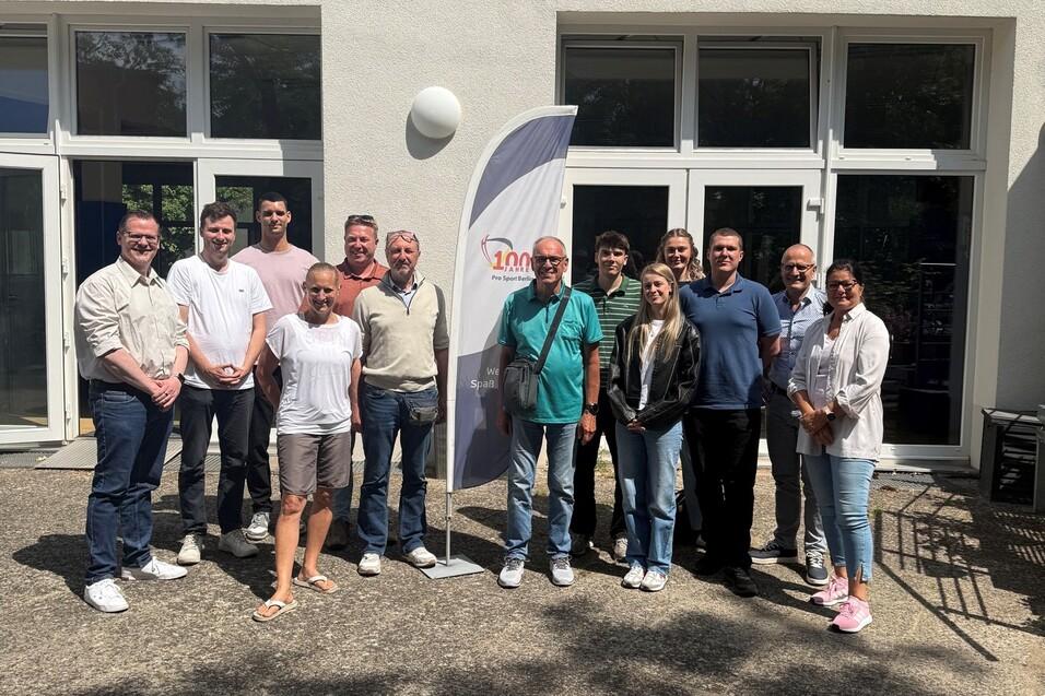 Gruppe von zw&ouml;lf Personen steht vor einem Geb&auml;ude, ein Banner mit der Aufschrift &bdquo;100&ldquo; ist sichtbar.