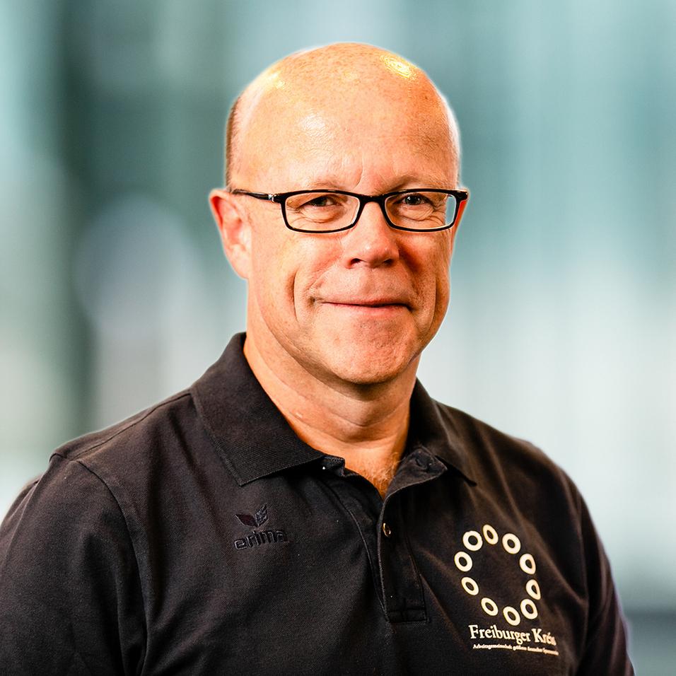 Porträt eines Mannes mit Glatze und Brille, der ein schwarzes Shirt mit Logo trägt, vor unscharfem Hintergrund.