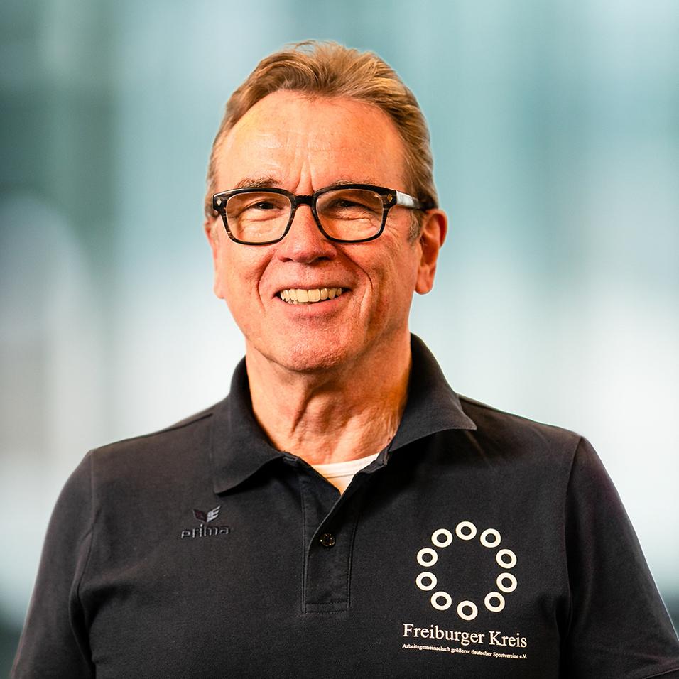 Portrait eines lächelnden Mannes in schwarzem Poloshirt mit Logo des Freiburger Kreises und Brille, unscharfer Hintergrund.