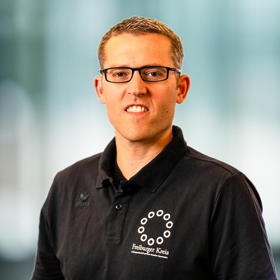 Porträt eines Mannes mit Brille, getragenem schwarzem Poloshirt mit Logo und neutralem Hintergrund.