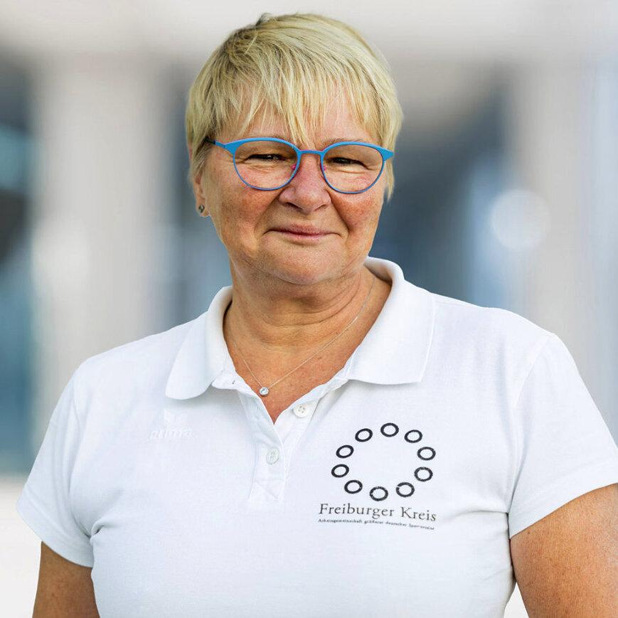Portrait einer lächelnden Frau mit blonden, kurzen Haaren und blauer Brille, in einem weißen Poloshirt mit Logo.