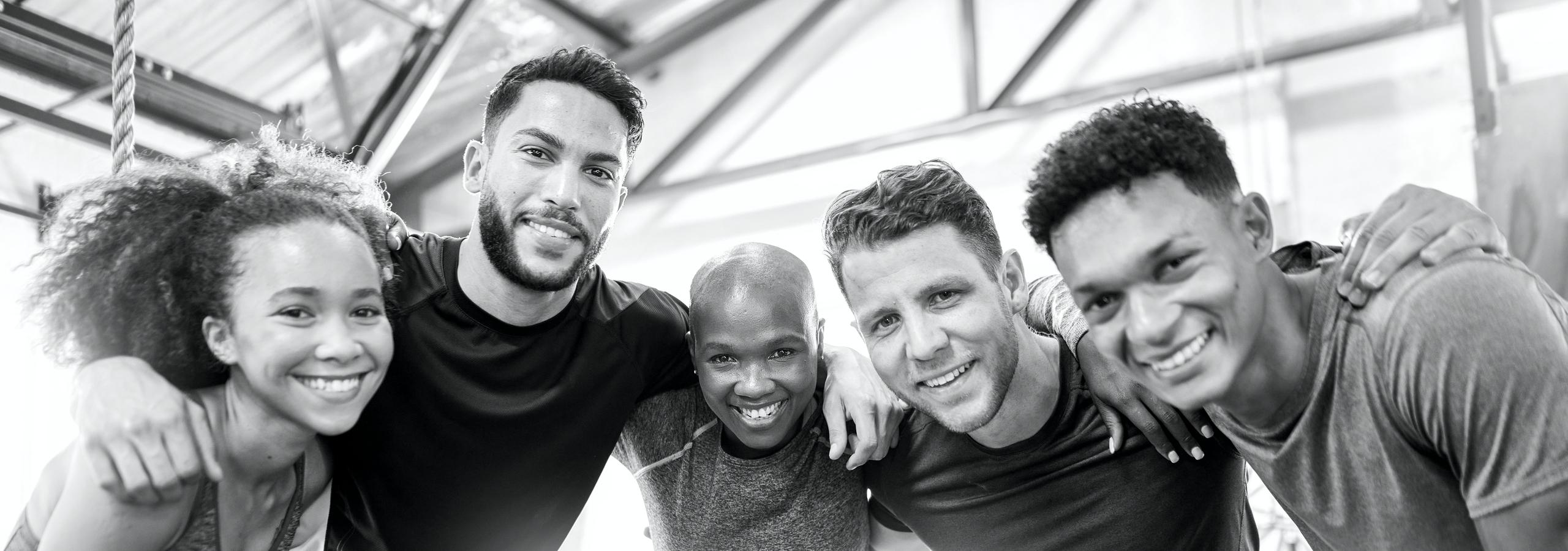 F&uuml;nf Sportler posieren l&auml;chelnd in einem Fitnessstudio, umarmt und in sportlicher Kleidung. Schwarz-Wei&szlig;-Aufnahme.