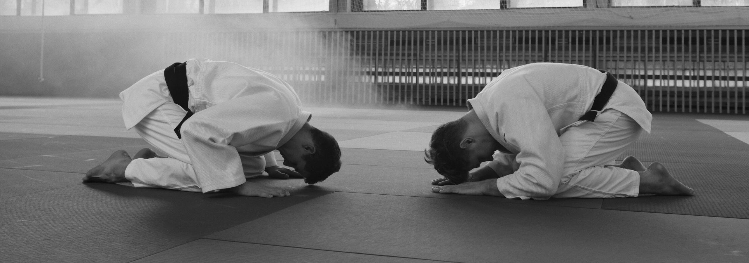 Zwei Judoka in wei&szlig;en Gis verbeugen sich respektvoll auf einer blauen Matte in einer Turnerhalle.