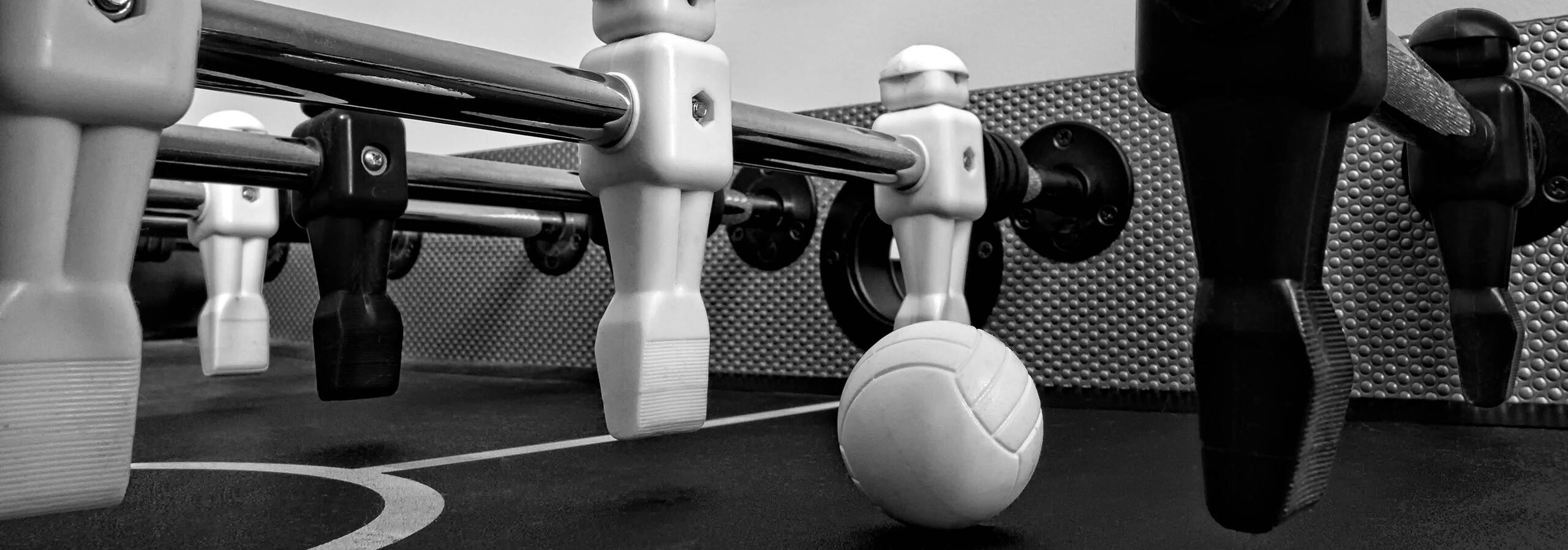 Nahaufnahme eines Tischfu&szlig;ballspiels mit wei&szlig;en und schwarzen Figuren sowie einem wei&szlig;en Ball auf einer Tischoberfl&auml;che.