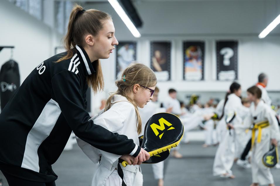 Trainerin unterstützt ein Kind beim Karate-Training mit Schläger in einem Übungsraum mit weiteren Kindern.